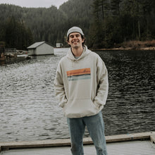 Load image into Gallery viewer, Model wearing sand coloured hooded sweatshirt with horizon design. Mountain Lake scenery in the background.