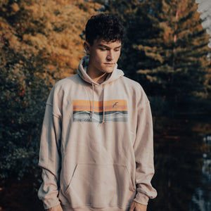 Model wearing sand coloured hooded sweatshirt with horizon design. Forest scenery in the background