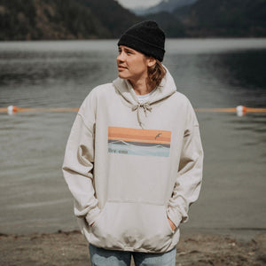 Model wearing sand coloured hooded sweatshirt with horizon design. Mountain Lake scenery in the background
