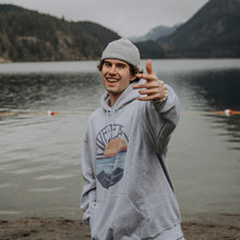 Load image into Gallery viewer, Model wearing grey hooded sweatshirt with Retro Sunset design. Mountain Lake scenery in the background.