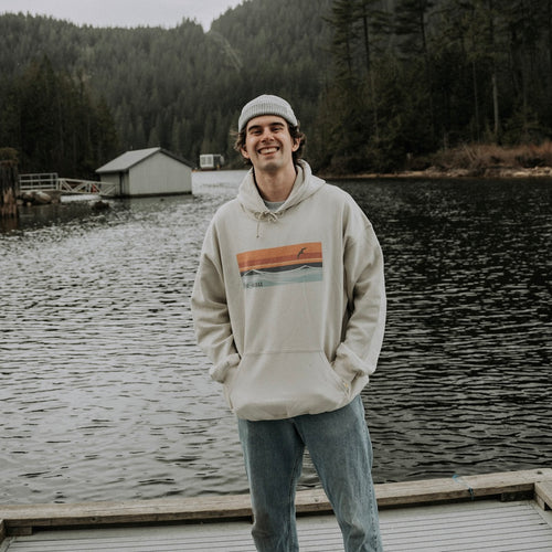 Model wearing sand coloured hooded sweatshirt with horizon design. Mountain Lake scenery in the background.